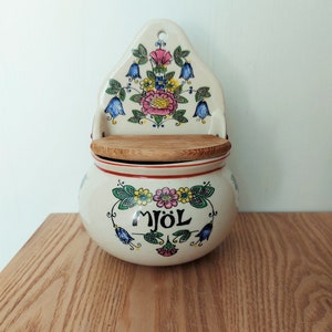Puede incluir: Un recipiente de harina de cerámica blanca de pared con tapa de madera. El recipiente está decorado con flores de colores y la palabra "MJOL" en letras negras.