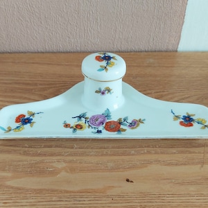 May include: A white porcelain inkwell with a floral design. The inkwell has a lid and a tray for holding pens. The floral design features pink, orange, yellow, and blue flowers.