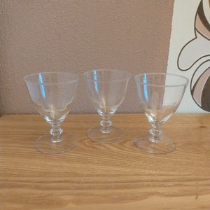 Peut inclure: Trois verres à pied transparents avec un bol arrondi et une tige décorative. Les verres sont disposés sur une surface en bois clair. L'arrière-plan présente un mur texturé et une partie d'un motif.