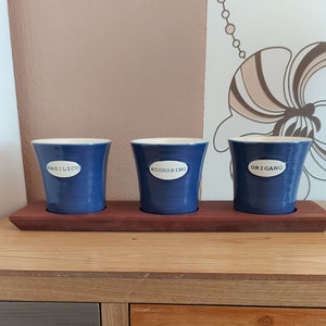 May include: Three blue ceramic herb pots with white interiors and labels that read "BASILICO", "ROSMARINO", and "ORIGANO". The pots sit on a dark wooden tray.
