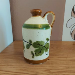 May include: A ceramic jug with a handle, featuring a speckled cream-colored body with a green band and brown accents. The jug is adorned with green leaf and vine designs. The base and top are brown.