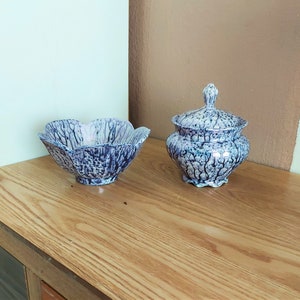 以下が含まれることがあります： 青と白の斑点模様の装飾用セラミックボウルと蓋付きジャーのセット。ボウルは花びらのような形をしており、ジャーは丸みを帯びたボディで、蓋には装飾的なつまみが付いています。木製の表面に置かれています。