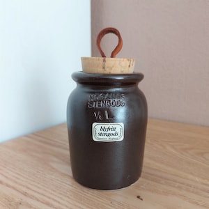Könnte beinhalten: Ein brauner Steinzeugtopf mit Korken und Lederschlaufe. Der Topf trägt die Prägung "HOGANAS STENGODS" und "1/2 L". Ein Aufkleber auf dem Topf lautet "blyfritt stengods stoneware Bleifrei".