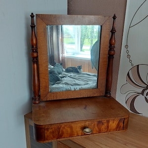 Peut inclure: Un miroir de coiffeuse en bois vintage avec un petit tiroir. Le miroir rectangulaire est encadré de bois brun clair, soutenu par deux montants verticaux ornés. Le tiroir a une poignée de couleur laiton. Le miroir reflète une pièce avec des chats.