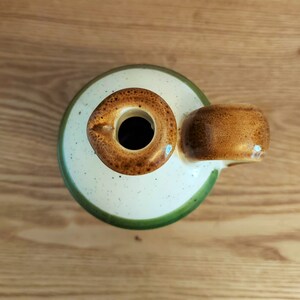 May include: A ceramic jug with a round opening and a handle. The jug has a speckled cream-colored body with a green rim and a brown handle and opening. The jug is sitting on a wooden surface.