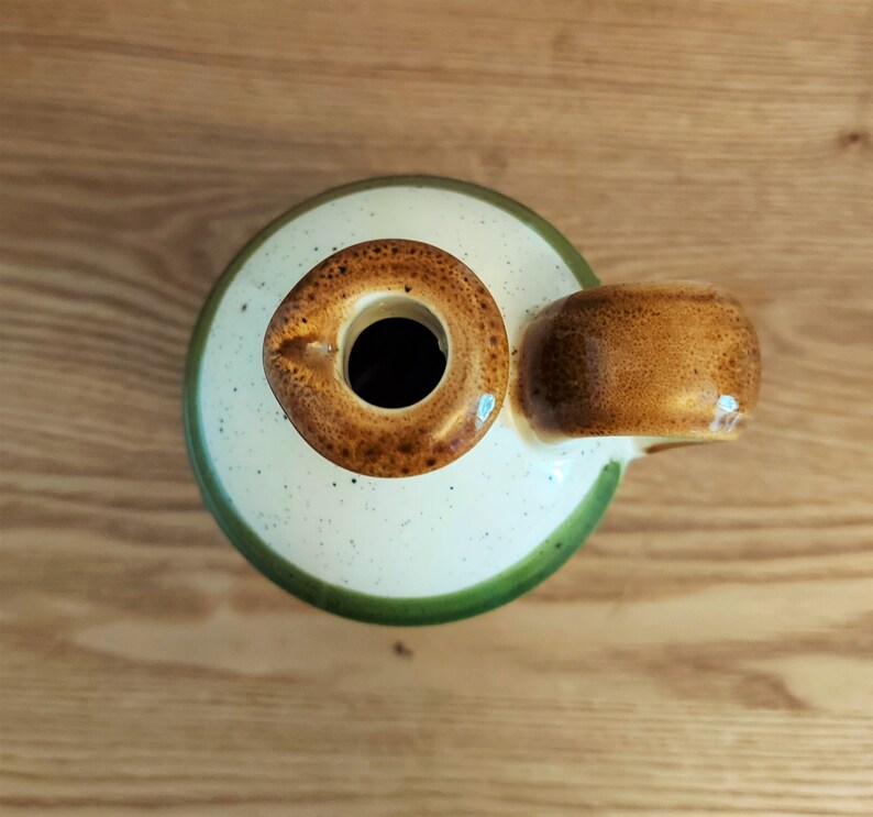 May include: A ceramic jug with a round opening and a handle. The jug has a speckled cream-colored body with a green rim and a brown handle and opening. The jug is sitting on a wooden surface.