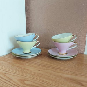Peut inclure: Un ensemble de quatre tasses à thé et soucoupes de couleur pastel. Les tasses sont empilées par paires, avec une tasse bleu clair sur une tasse vert pâle, et une tasse rose sur une tasse jaune. Les soucoupes sont blanches avec une bordure dorée.