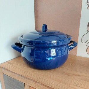 Op de afbeelding: Een blauwe emaille pan met een deksel. De pan heeft twee handvatten en een ronde vorm. De pan staat op een houten oppervlak.