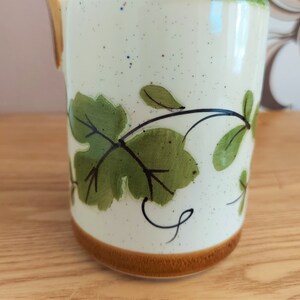 May include: Cream-colored ceramic mug with a brown base and handle. The mug features a hand-painted design of green leaves and vines. The mug has a speckled finish and a simple, elegant design.