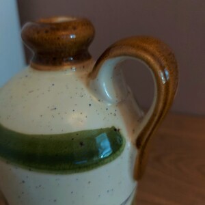 May include: A ceramic pitcher with a cream-colored body speckled with blue and brown dots. It features a brown handle and rim, with green leaf and vine designs. The pitcher has a classic, rustic aesthetic.
