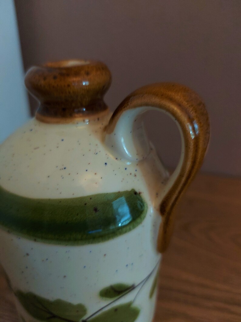 May include: A ceramic pitcher with a cream-colored body speckled with blue and brown dots. It features a brown handle and rim, with green leaf and vine designs. The pitcher has a classic, rustic aesthetic.