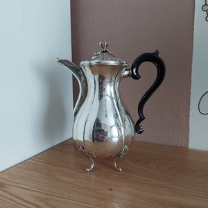 Op de afbeelding: Een zilveren theepot met een zwarte handgreep en drie poten. De theepot heeft een geribbeld lichaam en een decoratief deksel.