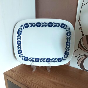 May include: A rectangular white serving tray with rounded corners. The tray has a blue floral border design. The tray is displayed on a clear plastic stand, highlighting its decorative pattern and shape.