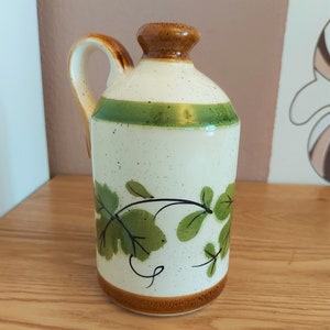 May include: A ceramic pitcher with a handle, featuring a cream-colored body speckled with brown dots. It has a brown rim and base, a green stripe, and green leaf and vine designs. The handle is brown with a cream interior.