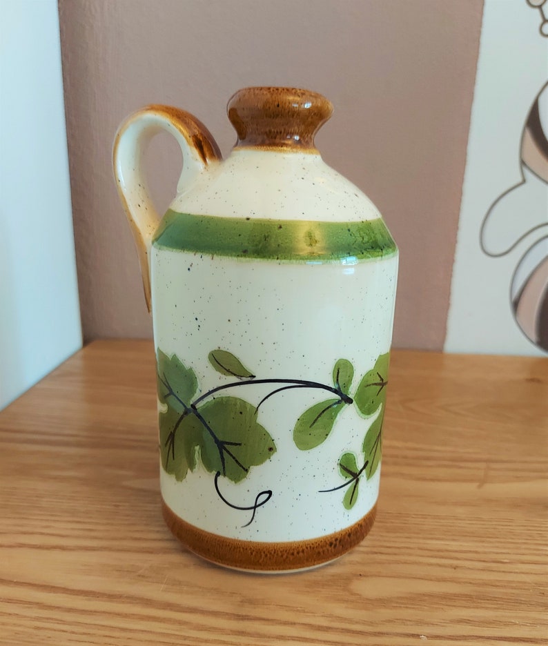 May include: A ceramic pitcher with a handle, featuring a cream-colored body speckled with brown dots. It has a brown rim and base, a green stripe, and green leaf and vine designs. The handle is brown with a cream interior.