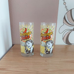 Peut inclure: Deux verres à boire transparents avec un design rétro "Orange Juice". Chaque verre présente une illustration vintage d'une femme tenant une bouteille, avec un motif à pois jaunes et un graphisme d'agrumes. Le texte "100% naturel" est également inclus.