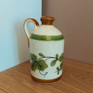May include: A ceramic pitcher with a handle, featuring a cream-colored body speckled with brown dots. It has a brown rim and base, a green stripe, and a brown top. Green leaf and vine design decorates the front.