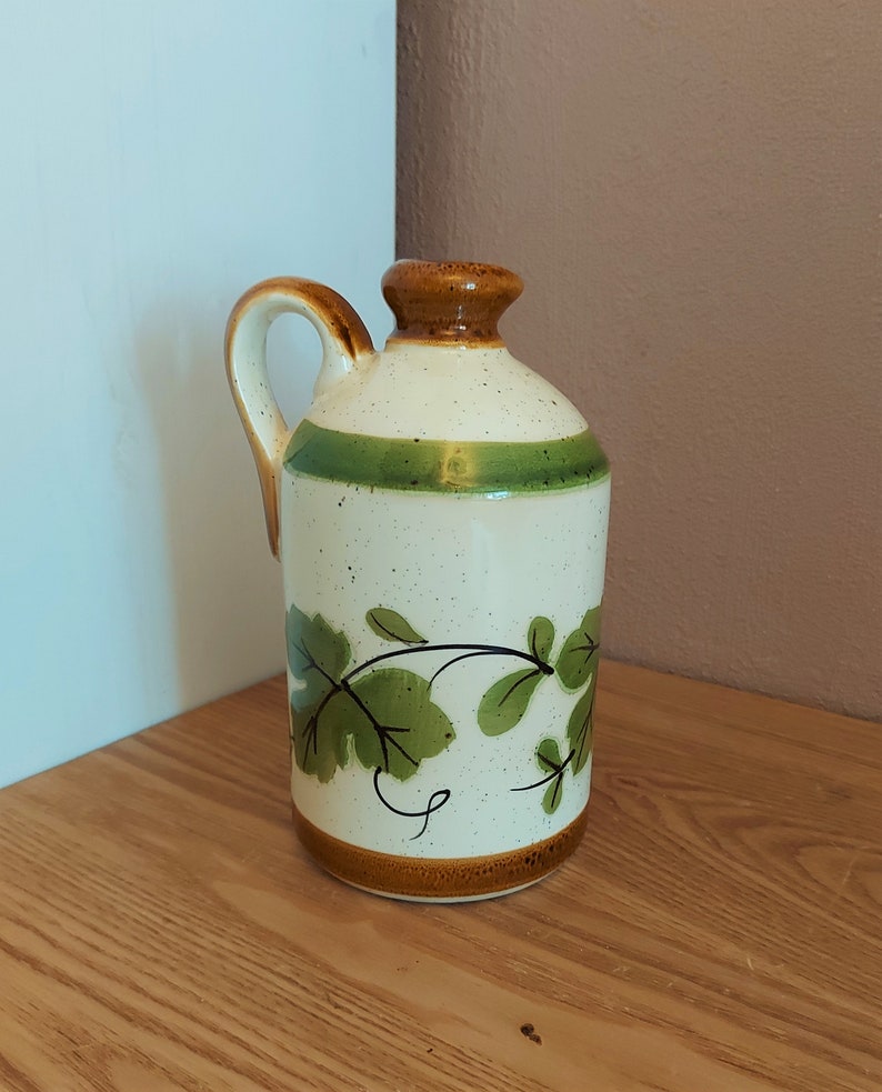 May include: A ceramic pitcher with a handle, featuring a cream-colored body speckled with brown dots. It has a brown rim and base, a green stripe, and a brown top. Green leaf and vine design decorates the front.