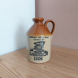 Puede incluir: Una jarra de cerámica marrón con asa. La jarra tiene una etiqueta que dice "Cornish Mead Wine Jugged By Jamaica Inn".