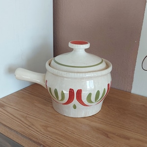 Puede incluir: Olla de cerámica blanca con tapa, con un diseño floral verde y rojo. La olla tiene un asa y una forma redondeada.