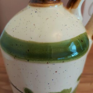 May include: A ceramic pitcher with a cream-colored body speckled with brown and blue dots. It features a wide green band and a brown top and handle. Green leaf designs are painted near the base, creating a rustic, handcrafted aesthetic.