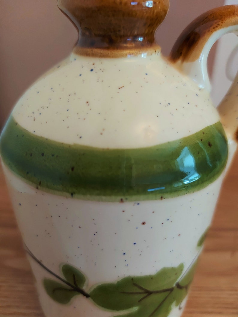 May include: A ceramic pitcher with a cream-colored body speckled with brown and blue dots. It features a wide green band and a brown top and handle. Green leaf designs are painted near the base, creating a rustic, handcrafted aesthetic.