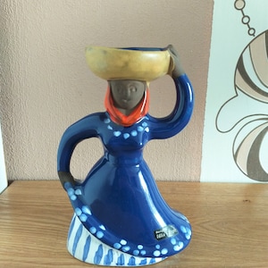 Może przedstawiać: Niebieska ceramiczna figurka kobiety w niebiesko-białej sukience, która nosi żółtą miskę na głowie. Figurka jest ozdobiona białymi kropkami i ma czerwoną chustkę wokół szyi. Figurka jest podpisana "Design Lilla Scott".
