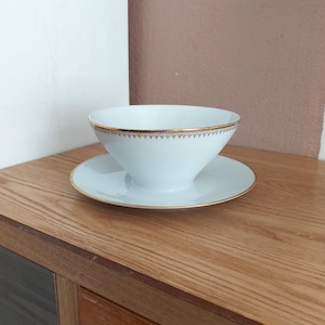 May include: A white porcelain gravy boat with a gold trim and a matching saucer. The boat has a wide, rounded base and a narrow spout.
