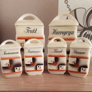 Könnte beinhalten: Satz von sechs vintage Keramikbehältern mit Deckeln. Die Behälter sind mit einem geometrischen Muster in Braun-, Orange- und Weißtönen verziert. Die Deckel haben handgeschriebene Etiketten in Schwarz, darunter "Frukt", "Havregryn", "Kanel", "Vitpeppar", "Kardamomma" und "Kryddpeppar".