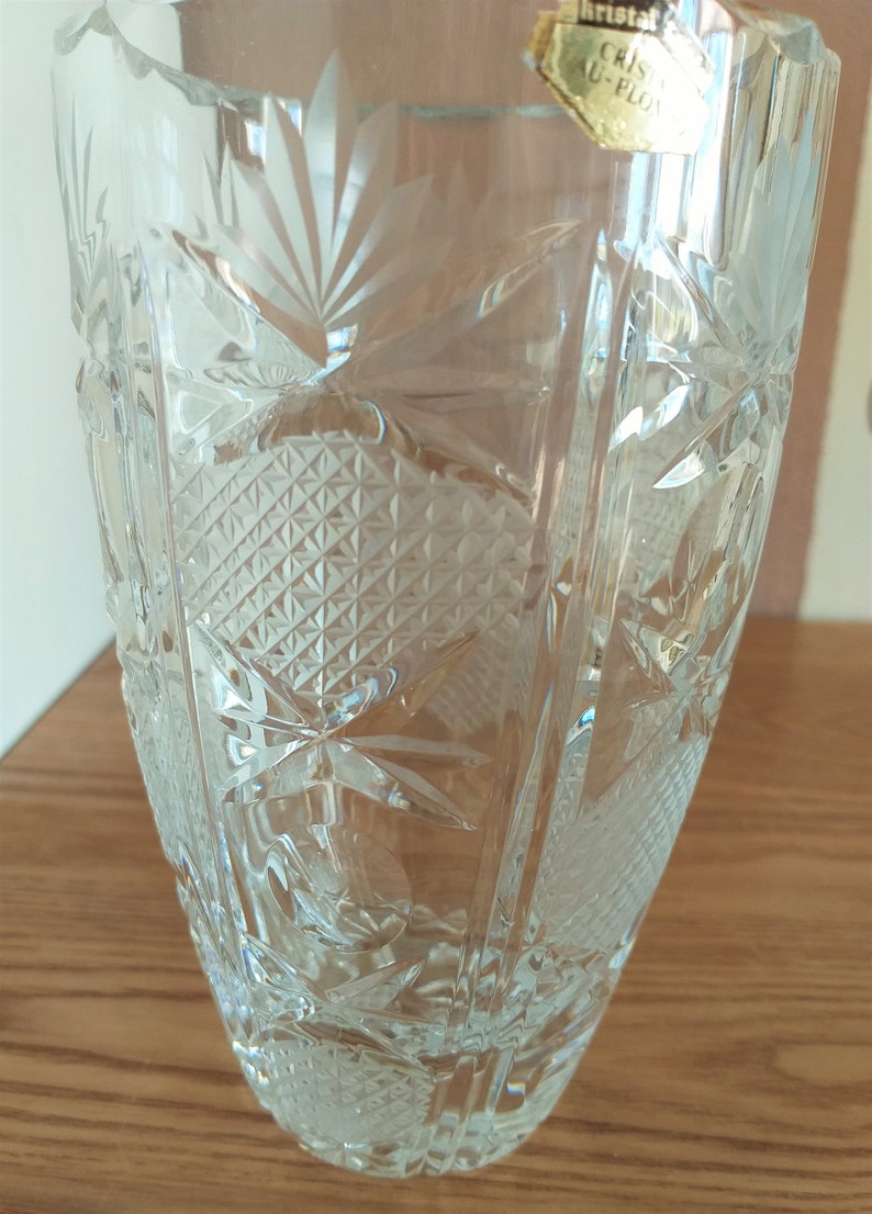 May include: A clear glass vase with a cut crystal design. The vase has a label that reads "Cristal Au-Ploy".