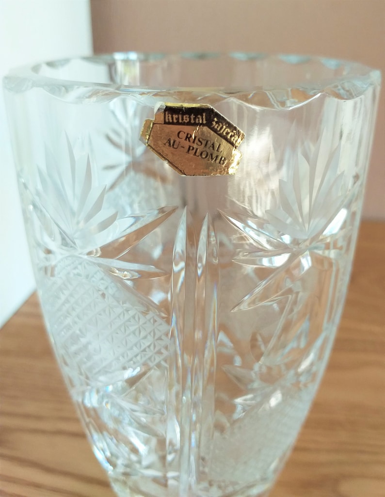 May include: A clear glass vase with a cut crystal design. The vase has a label that reads "Kristal Sajica Cristal Au-Plomb".