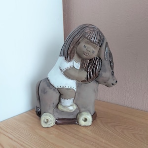 May include: A ceramic figurine of a child riding a donkey. The child wears a white dress, and the donkey is grey. The figurine is displayed on a wooden surface.