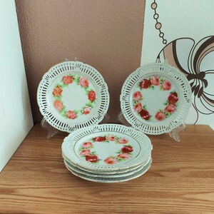 Peut inclure: Ensemble de quatre assiettes en porcelaine blanche avec un motif floral délicat représentant des roses roses et rouges. Les assiettes ont un bord festonné et un motif en dentelle autour du bord.