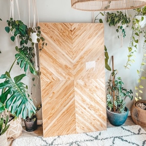 May include: A large wooden wall panel with a chevron pattern. The panel is light brown and has a natural wood finish. The panel is surrounded by plants and a white rug.
