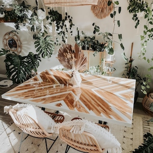 May include: A rectangular dining table with a glossy white and natural wood geometric inlay pattern, paired with two curved rattan chairs featuring white fringed cushions, positioned on a textured cream rug in a room filled with various green plants.