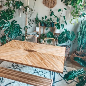 May include: A rectangular wooden dining table with a chevron-patterned surface and thin metal legs, accompanied by a matching slatted wooden bench. The table features a light brown wood grain finish and is positioned in a room filled with various green houseplants.