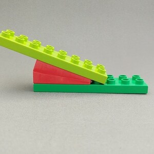 May include: A green and red plastic toy seesaw made of Lego bricks. The seesaw is made of three pieces: a green base, a red wedge, and a long green piece with studs on top.
