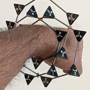 May include: A black metal sculpture of a geometric shape made of triangular pieces with numbers on each side. The sculpture is held in a person's hand.