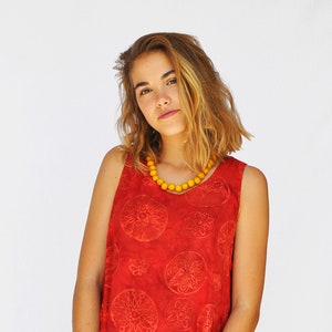 Può includere: Un top rosso senza maniche con un design a strati e un motivo circolare ripetuto. Il top è abbinato a pantaloni bianchi e una collana di perline gialle. Un braccialetto nero è indossato al polso.