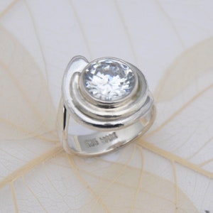 Peut inclure: Une bague en argent avec une grande pierre ronde transparente sertie dans un motif en spirale. La bague est marquée "925" à l'intérieur de la bande.