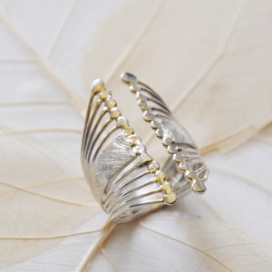 Könnte beinhalten: Ein silberfarbener Ring mit einem einzigartigen Design. Der Ring hat eine fächerartige Struktur mit mehreren vertikalen Linien und kleinen, abgerundeten Details an den Kanten. Der Ring ist vor einem hellen Hintergrund platziert.
