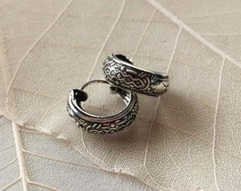 May include: A pair of silver hoop earrings with a detailed, intricate design. The earrings are on a light brown background.