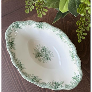 May include: Oval-shaped, antique white serving dish with a green floral pattern and scalloped edges. The dish features a central floral bouquet and decorative border. A classic piece for serving food.