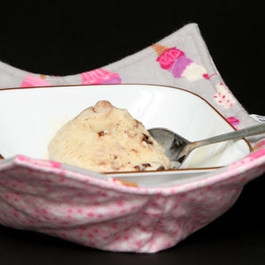 May include: A white rectangular bowl with ice cream and a spoon rests inside a gray and pink fabric bowl cozy. The cozy has a pink star pattern and a gray border with ice cream cone designs.
