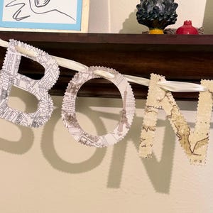 Puede incluir: Una pancarta decorativa que deletrea "BON" con letras individuales recortadas de papel. Las letras están unidas a una cinta blanca. Las letras están decoradas con mapas y diseños de ciudades.