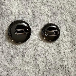 Op de afbeelding: Twee zwarte, ronde knopen met een glanzende afwerking. Elke knoop heeft een zilveren paperclip in het midden. De knopen zijn van verschillende grootte en liggen op een grijze, getextureerde ondergrond.