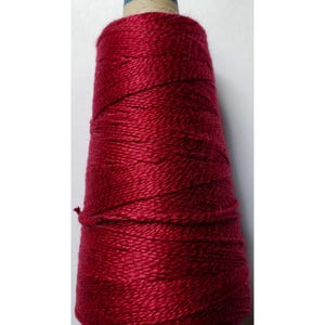 May include: A conical spool of deep red yarn wound tightly around a cardboard core. The yarn features a smooth, slightly lustrous texture with visible individual strands wrapped in a consistent diagonal pattern from the base to the top.