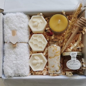 May include: A white gift box filled with a white towel, four honey-scented soaps shaped like honeycombs with bees, a small jar of honey, a small bottle of bath salts, a small candle, and a wooden honey dipper.