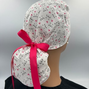 May include: White scrub cap with a repeating pattern of black cat faces and pink hearts. A bright pink ribbon is tied in a bow at the back. The cap is worn by a person wearing a black top.