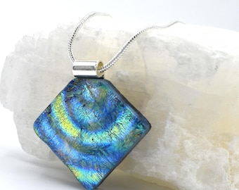 May include: A dichroic glass pendant necklace with a square, diamond-shaped pendant. The pendant features swirling patterns of blue, green, and yellow. The necklace has a silver-colored chain and a silver-colored metal bail.
